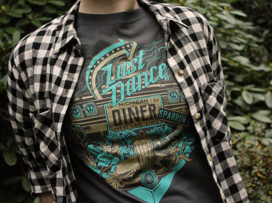 An illustrated typography graphic tshirt that reads "Last Dance Diner" and features iconography from the book Sparrow Hill Road by Seanan McGuire. The shirt is charcoal gray with teal and gold ink. The illustration includes a license plate, roses, a burger and milkshake diner meal, and road sign. 