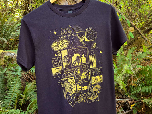 A black t-shirt with a metallic gold graphic screen print of a sci-fi astronaut and space-themed illustrations, hanging on a wooden hanger against a lush green forest background.