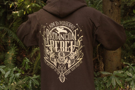 A person wearing a dark brown sweatshirt featuring a large distressed gold graphic print with the text 'I AM TO MISBEHAVE BROWNCOAT REBELLION' and illustrated details.
