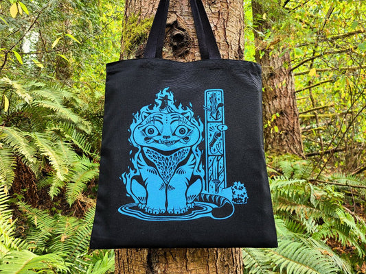 Black tote bag with blue graphic design of Derpy Tiger hanging on a tree in a forest setting