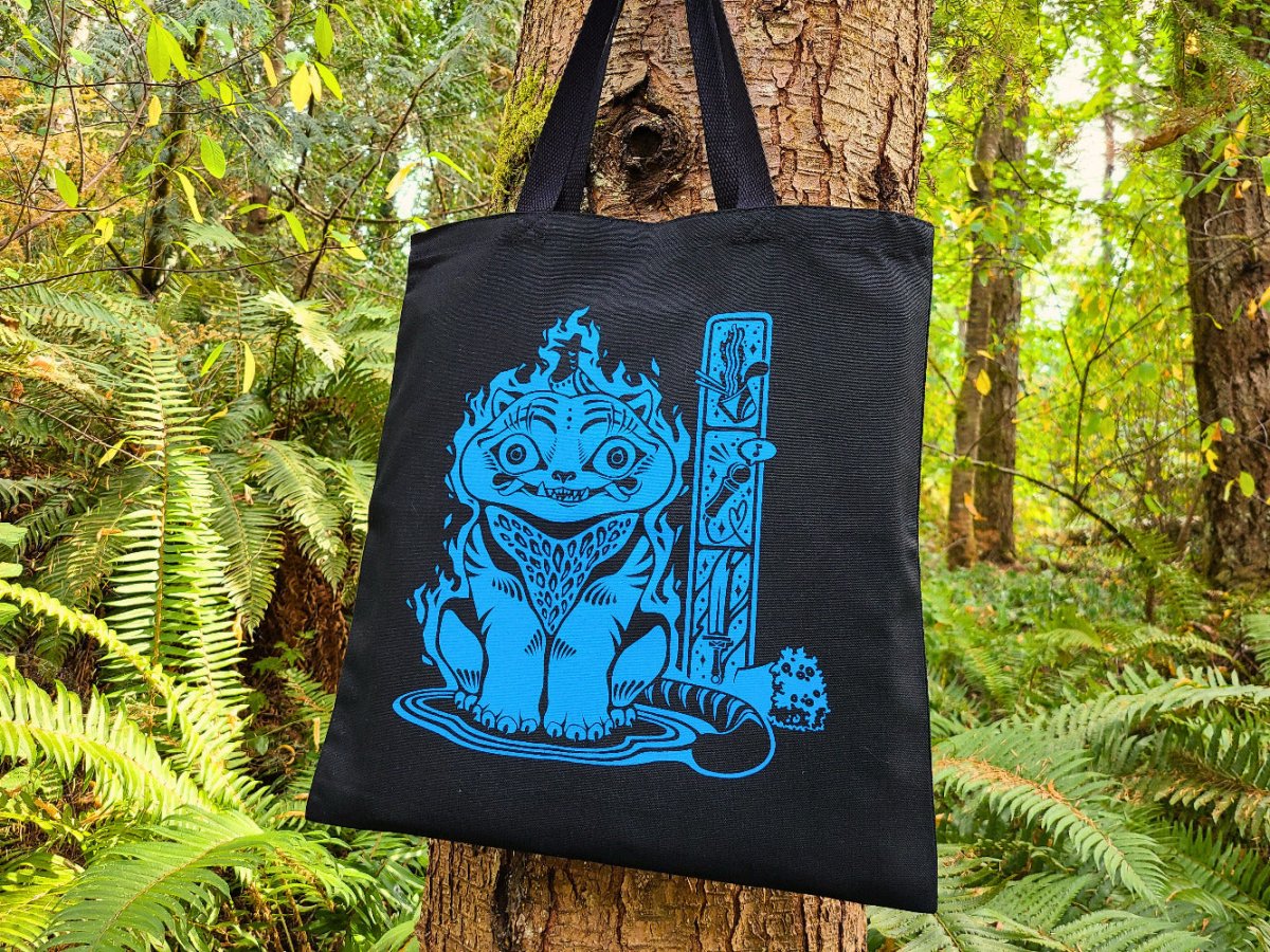 Black tote bag with blue graphic of Derpy Tiger hanging on a tree in a forest