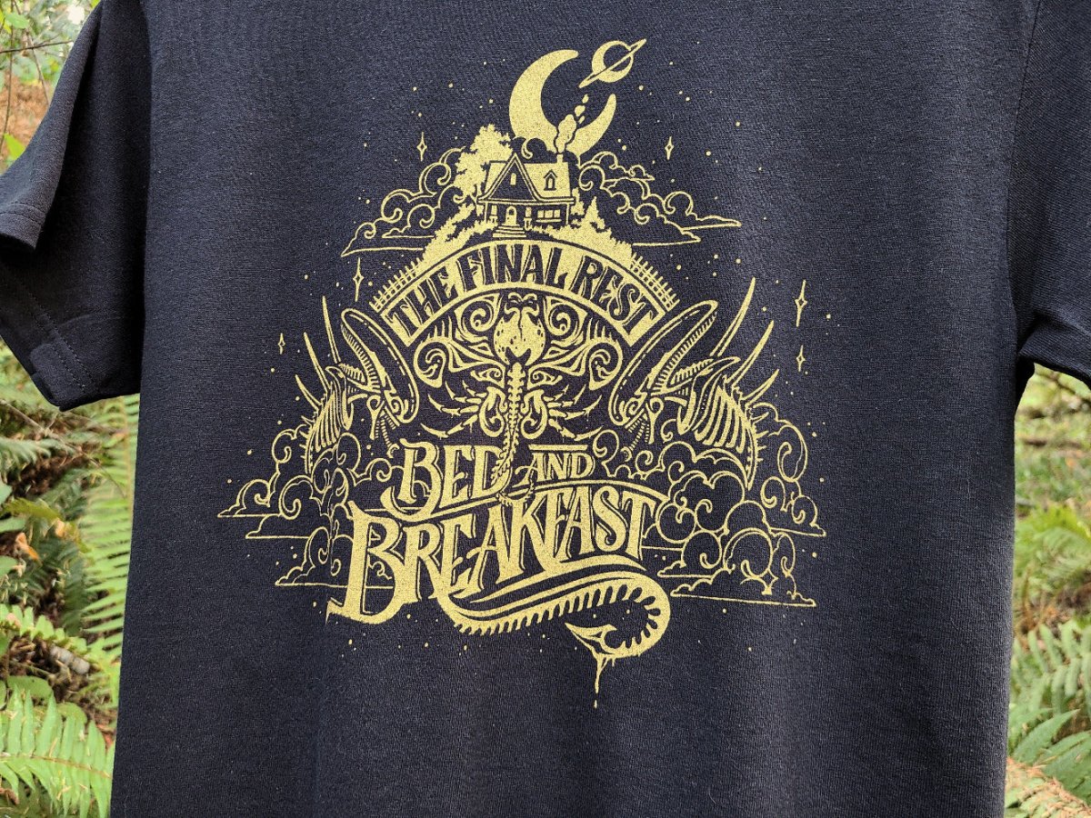 Black t-shirt with gold graphic design featuring an Alien scifi design, against a natural background.