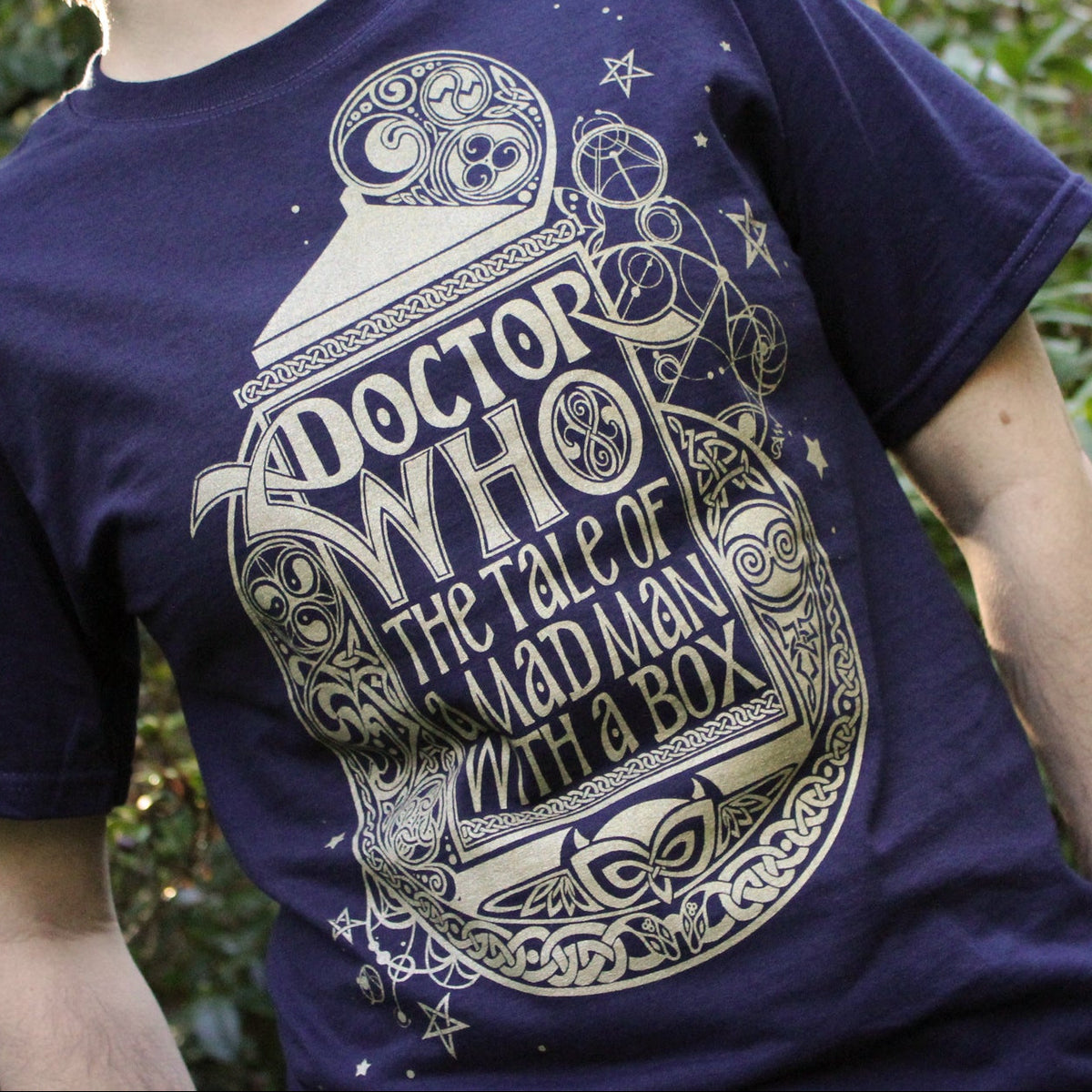A person wearing a navy blue t-shirt with a large gold Doctor Who graphic on the front, set against a blurred green outdoor background.