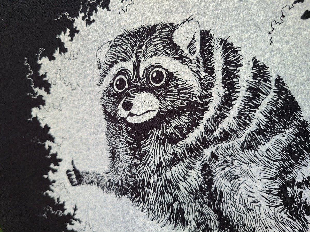 A close-up of a black and white graphic print featuring a detailed sketch of a raccoon's face and upper body, with textured fur lines and a dark background.