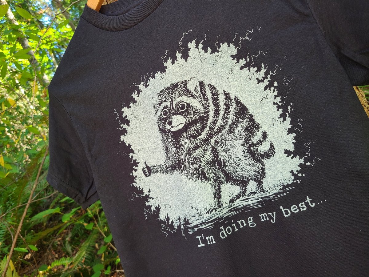 A close-up of a black t-shirt featuring a distressed white graphic of a raccoon giving a thumbs-up with the text 'I'm doing my best.