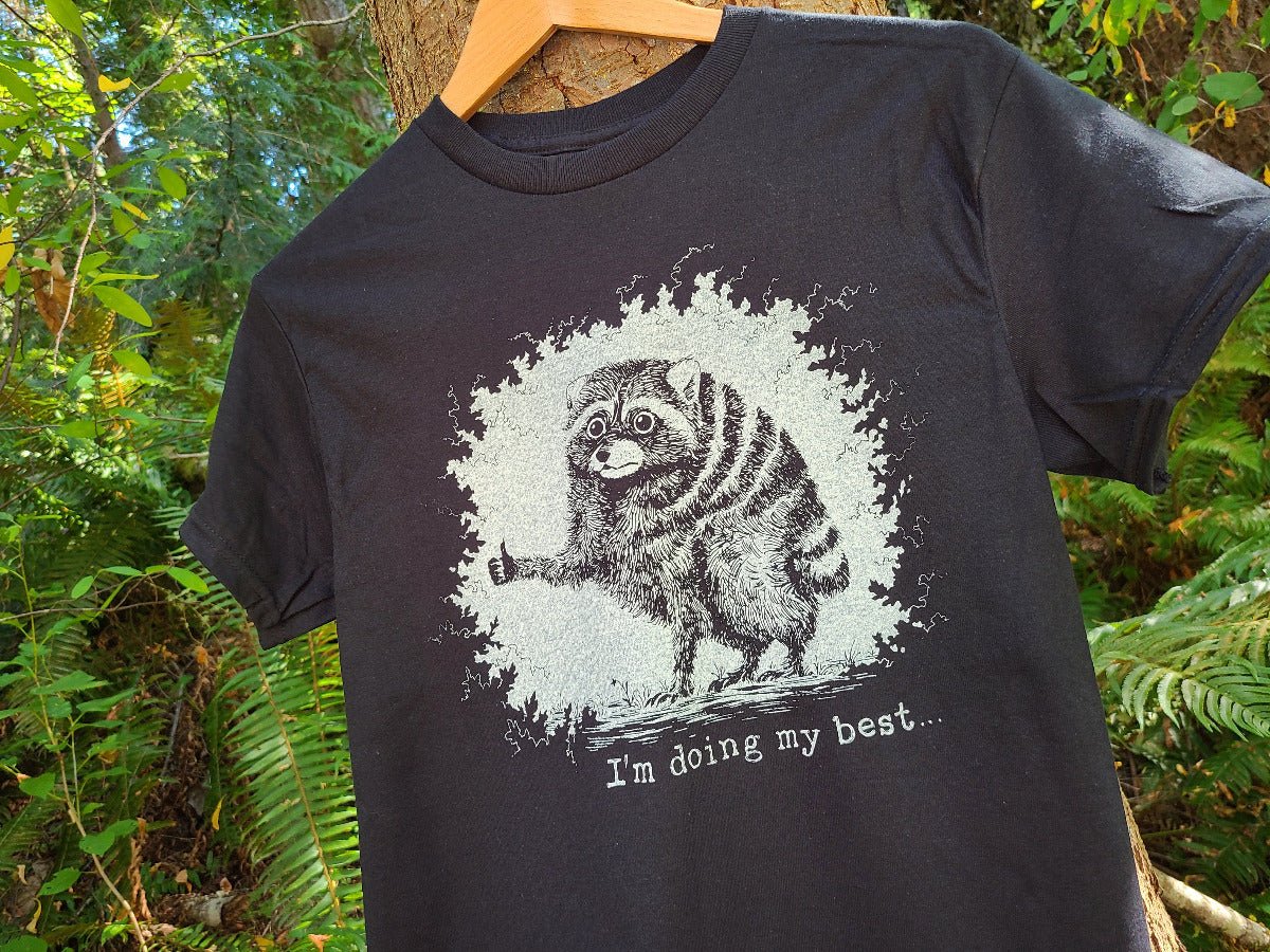 A black graphic t-shirt hanging on a wooden hanger against a lush green forest background. The shirt features a distressed white graphic of a raccoon giving  a thumbs up, with the text 'I'm doing my best.