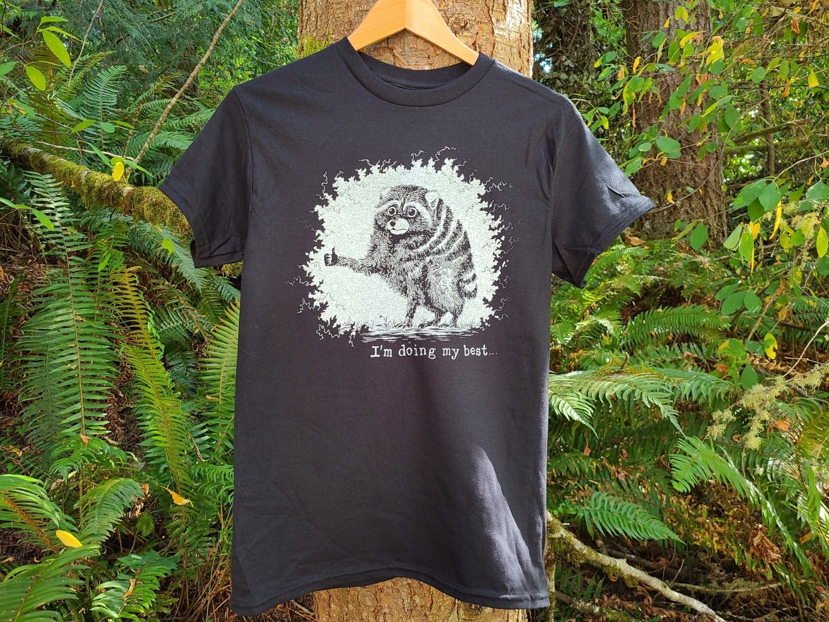 A black graphic t-shirt hanging on a wooden hanger against a lush green forest background. The shirt features a white distressed graphic of a raccoon giving a thumbs up and the text 'I'm doing my best.