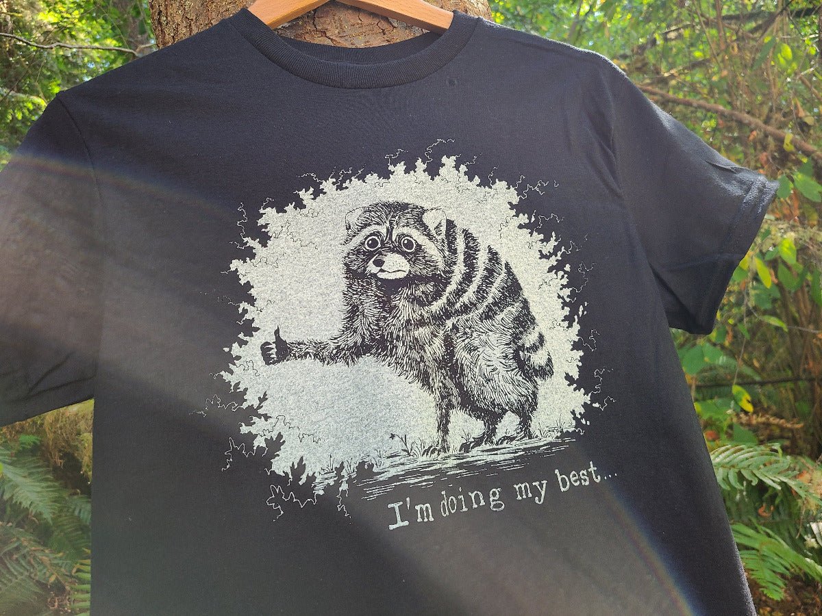 A black short-sleeve t-shirt hanging on a wooden hanger outdoors against a tree trunk and green foliage background. The shirt features a distressed white graphic of a raccoon giving a thumbs-up inside a splattered circle, with the text 'I'm doing my best.