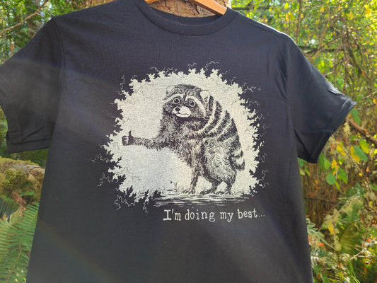 A black short-sleeve graphic t-shirt hanging on a wooden hanger outdoors against a background of green foliage. The shirt features a white distressed graphic of Jimothy the raccoon giving a thumbs-up with the text 'I'm doing my best.