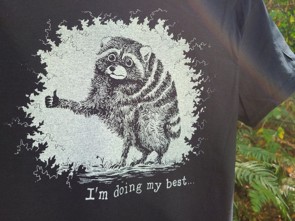 A close-up of a black t-shirt featuring a humorous graphic of a raccoon giving a thumbs-up inside a white circle, with the text 'I'm doing my best.