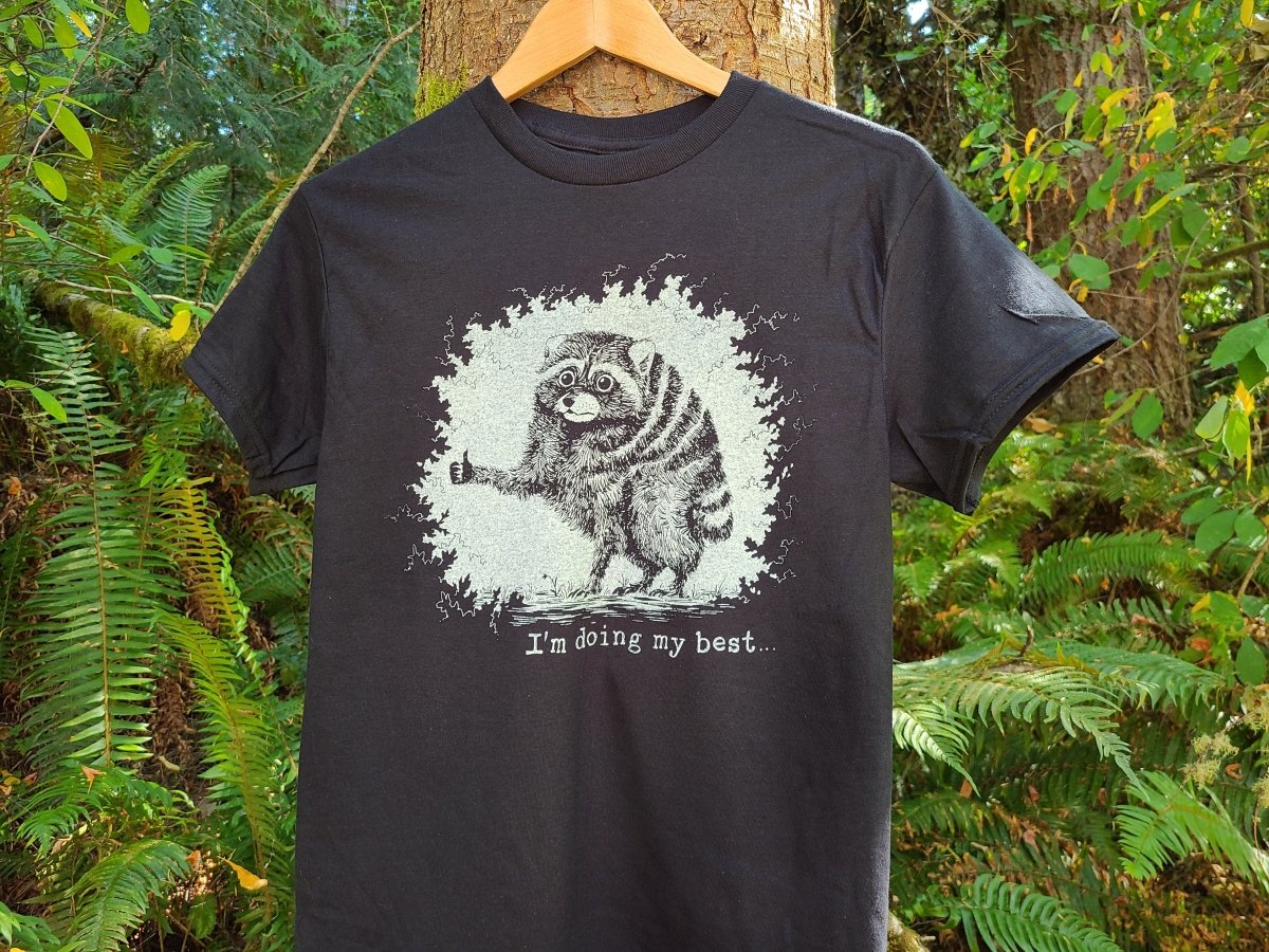 A black t-shirt hanging on a wooden hanger against a lush green forest background. The shirt features a distressed white graphic of a raccoon giving a thumbs up, with the text 'I'm doing my best.