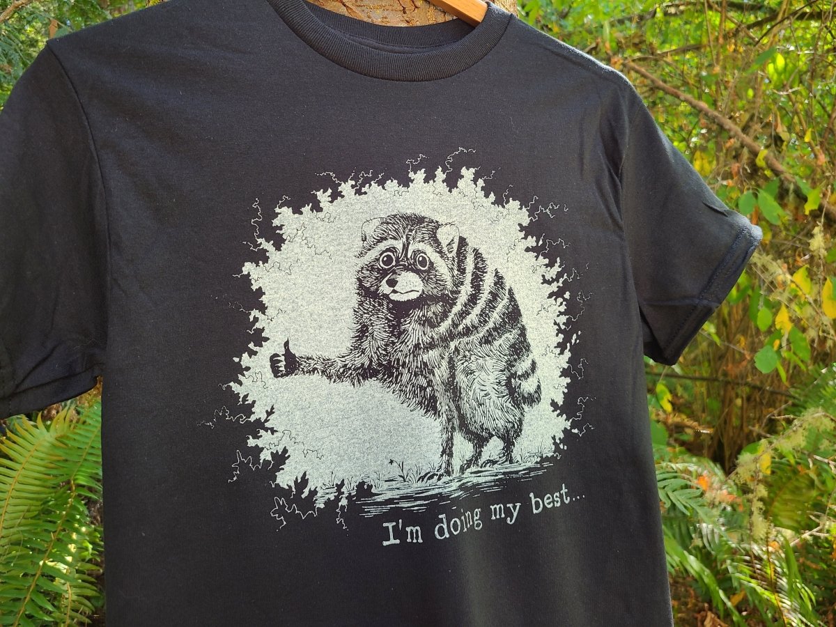 A black short-sleeve t-shirt hanging on a wooden hanger outdoors, featuring a distressed white graphic of a raccoon giving a thumbs-up with the text 'I'm doing my best.