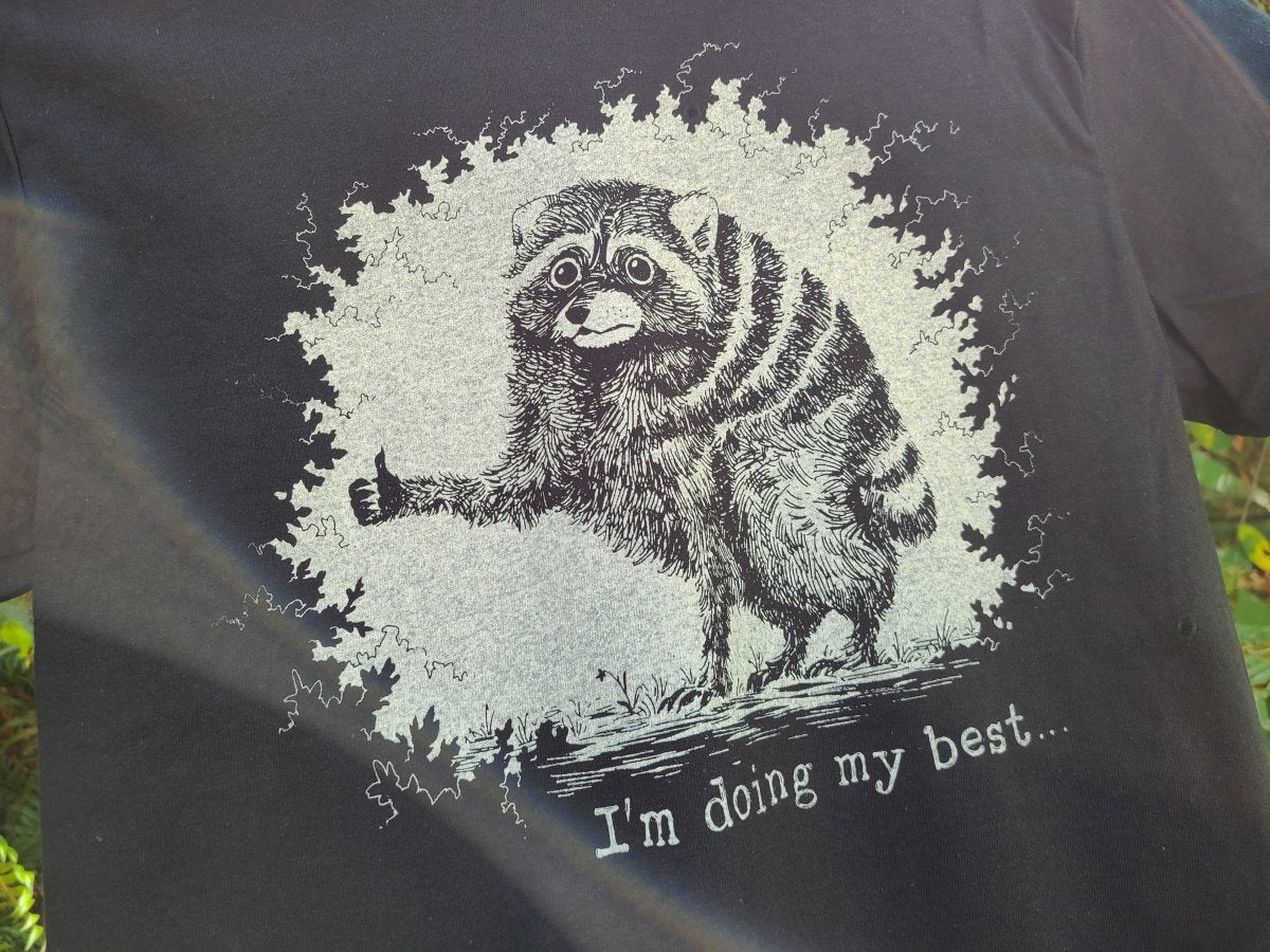A close-up of a black T-shirt featuring a humorous graphic of a raccoon giving a thumbs-up, with the text 'I'm doing my best.