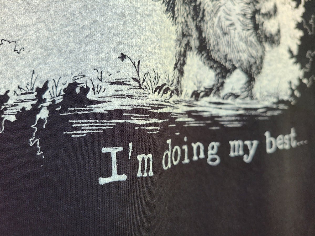 Close-up of a black graphic t-shirt featuring a black and white illustration of a raccoon standing in shallow water, with the text 'I'm doing my best.