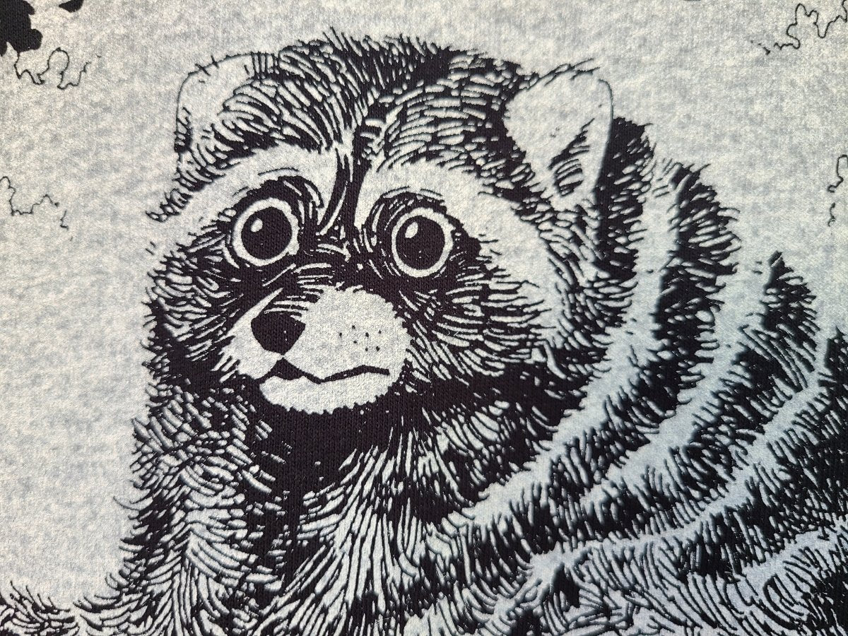 A close-up of a black and white graphic print depicting a raccoon's face and fur, set against a light gray textured background.