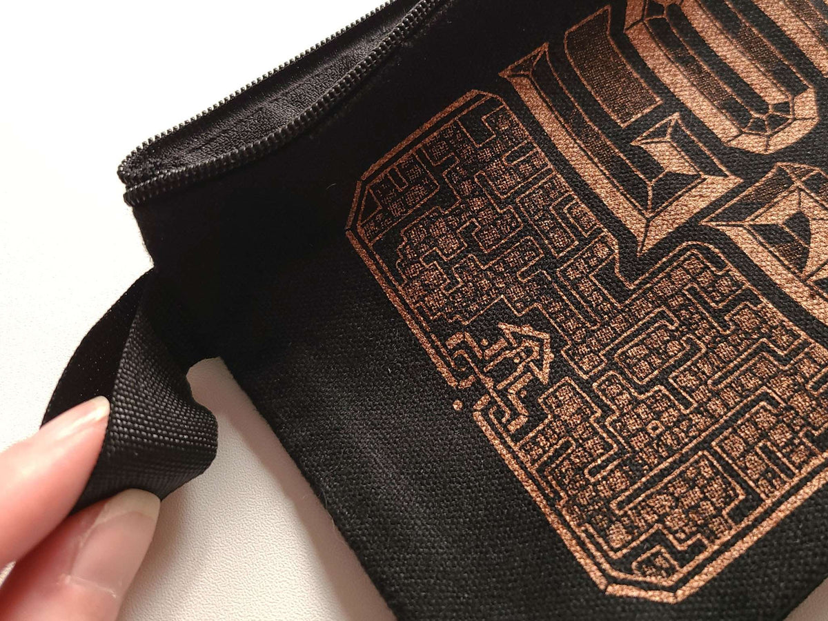 A close-up of a black canvas zipper pouch with a copper-toned geometric graphic print, held by a hand on a white background.