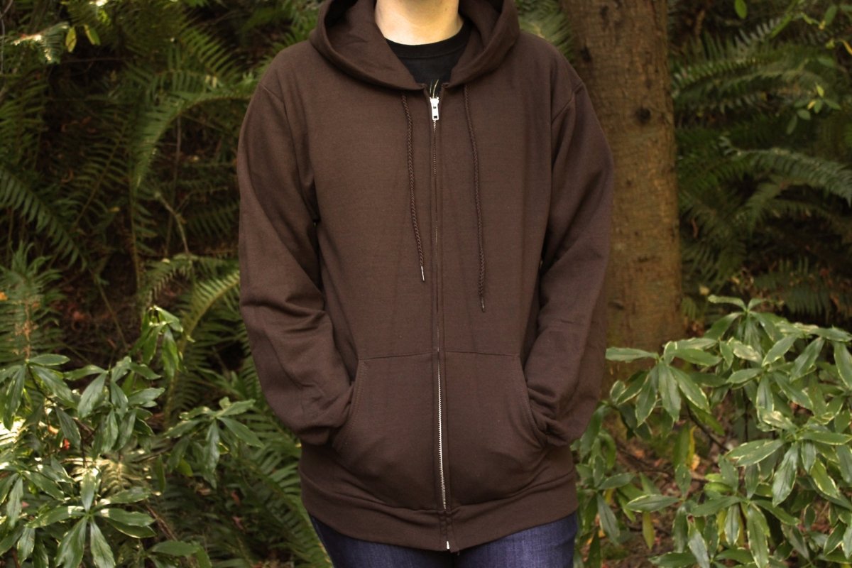 A person wearing a dark brown full-zip hoodie with drawstrings and kangaroo pockets, standing outdoors in front of a backdrop of green foliage and trees.