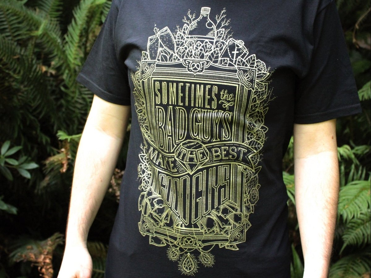Person wearing a black t-shirt with gold graphic design and text in a forest setting