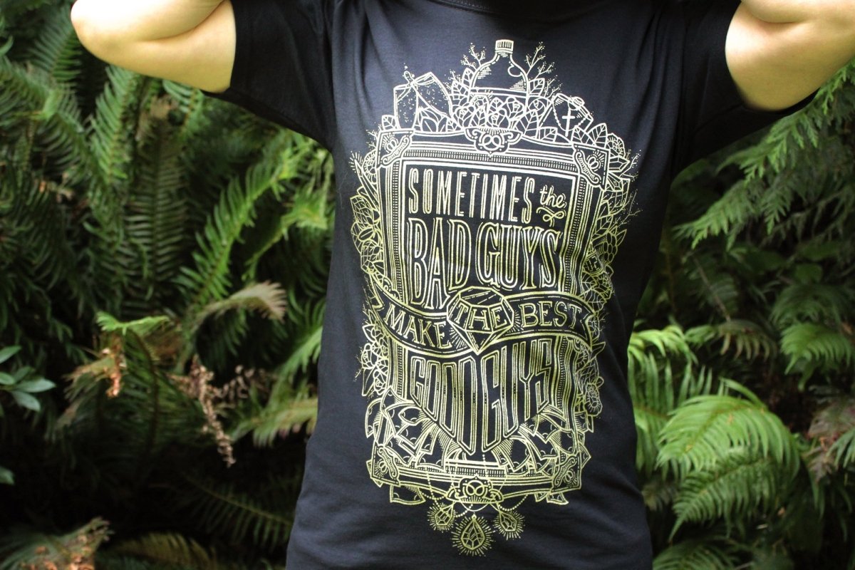 Person wearing a black t-shirt with gold graphic and text in front of green foliage