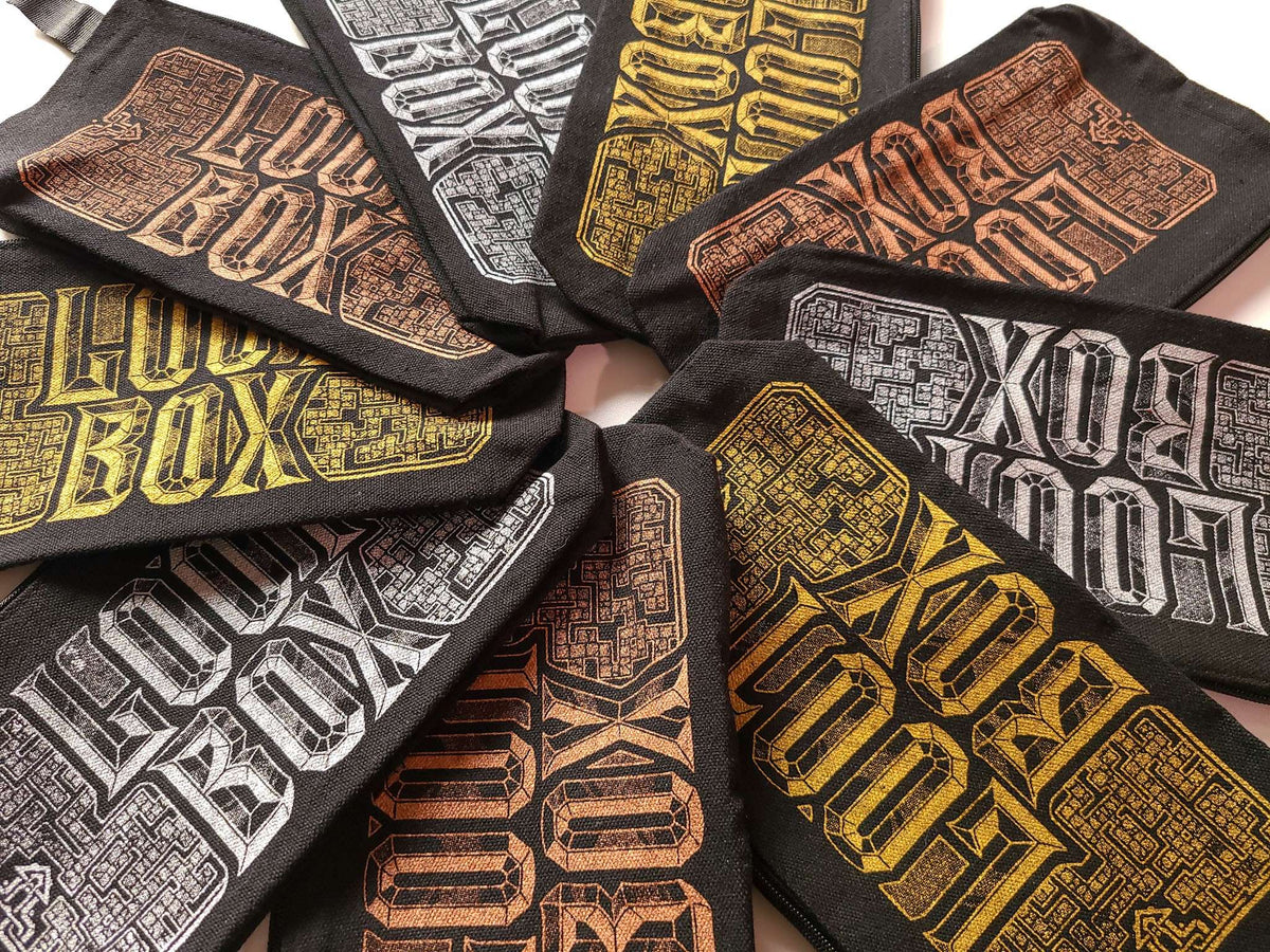 A close-up of several black fabric accessory bags arranged in a circle, each printed with the words 'LOOT BOX' in large stylized lettering. The lettering appears in gold, copper, and silver tones on the black fabric backgrounds.